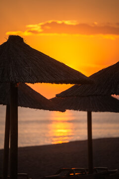 Straw sunshades on an empty sandy beach at sunset with golden sunlight reflecting on the sea