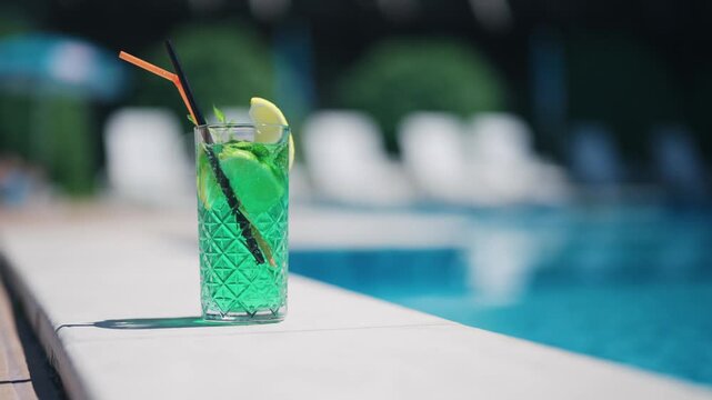 A green coctail drink with a straw in it is sitting on a ledge next to a pool. The drink is in a tall glass and has a lime wedge in it. Concept of relaxation and leisure