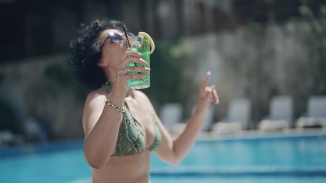A woman is dancing in slow motion by a pool with a drink in her hand. She is smiling and she is enjoying herself