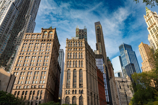A striking upward view of the mix of historic and modern buildings in downtown Manhattan, New York, featuring intricate stone facades alongside towering contemporary skyscrapers.