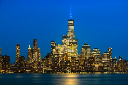 An illuminated view of downtown Manhattan glowing at dusk with city lights reflecting on the Hudson river in New York City.