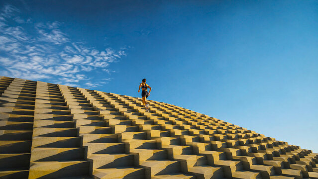 Athletic woman running up monumental geometric concrete stairs under a blue sky, ideal for sportswear, fitness, wellness, lifestyle and motivational advertising campaigns.