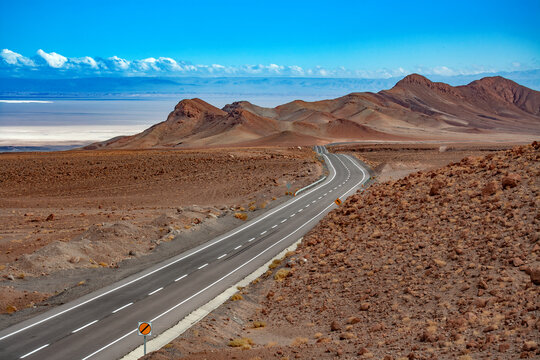 A long remote road traversing the arid landscape of the Atacama Desert in northern Chile, with the vast Salar de Atacama salt flat stretching across the distant background behind rocky hills.