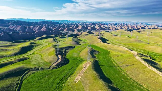 Aerial view of rolling green hills and red-brown Danxia formations with dirt tracks, transmission pylons, blue sky, clouds, and bright sunlight.