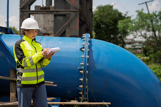 Focused professional female civil engineer holding a clipboard while inspecting large water gate infrastructure for flood control and environmental safety during an active outdoor operation.