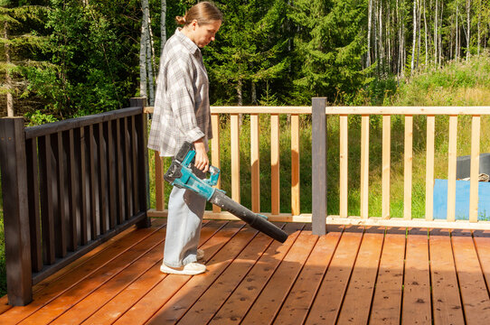 Woman cleaning a stained wooden deck with a cordless leaf blower, removing dust and debris during outdoor home maintenance and DIY work.