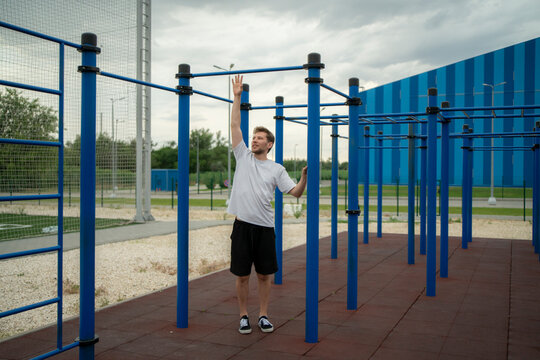 Man stands at outdoor fitness area with blue bars and gray sky, preparing for workout while looking around