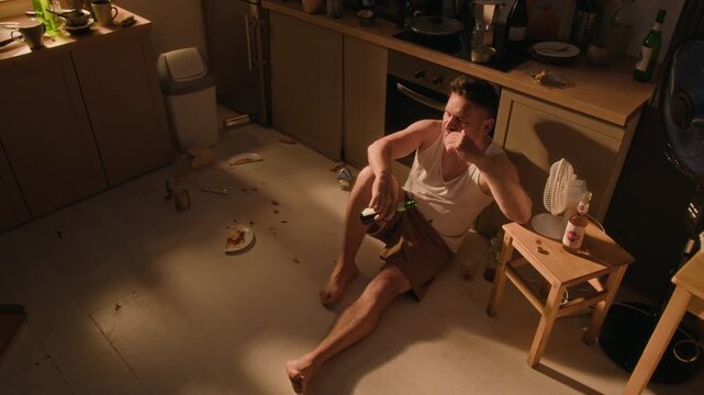 Depressed young adult Caucasian man sitting on messy floor drinking beer and holding head in high angle view. Suitable for addiction recovery and mental health campaigns