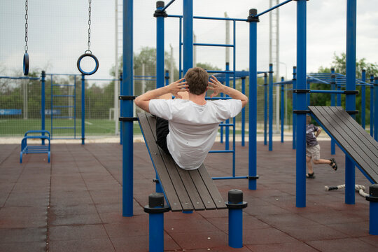 A man performs an ab workout on a bench in a fitness area while a child plays nearby in a park
