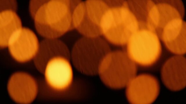 Blurred warm orange bokeh lights glow softly against a dark black background, with one bright circular light gradually growing larger and more intense over time throughout.