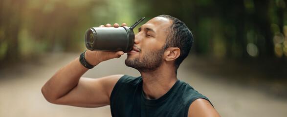 Fototapeta premium Happy Young Man Enjoying Hot Beverage From Black Thermos While Relaxing Outdoors, Focusing On His Wellbeing During Fitness Activities In A Natural Setting.