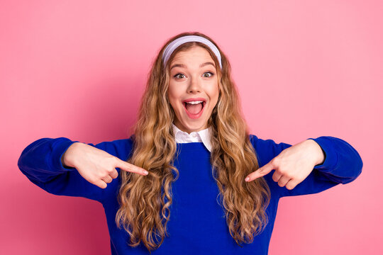 Young stylish millennial woman in blue sweater with headband smiles and points at herself against pink background