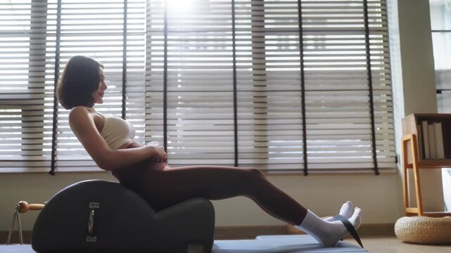 Woman practicing Pilates on a spine corrector, legs extended upward in a controlled stretch, bright minimalist studio with natural light and green plants in the background.	