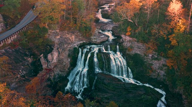 Curving roadway climbs through forested mountains beside rushing waterfall and colorful foliage. Scenic landscape offers peaceful atmosphere for driving and sightseeing.
