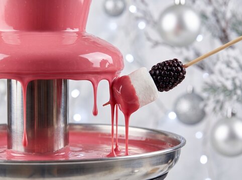 Pink ruby chocolate fountain cascading over marshmallow and blackberry skewer with silver Christmas lights