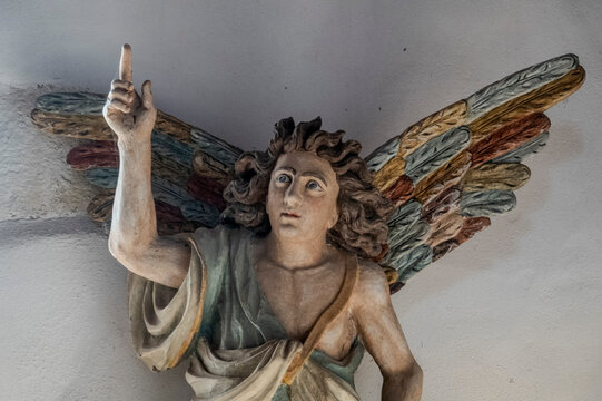 A carved wooden sculpture of an angel with colorful wings points upward against a wall.