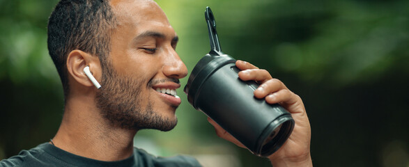 Fototapeta premium Smiling Young Arab Man With Light Brown Skin Enjoying A Cup Of Coffee While Listening To Music With Wireless Earbuds In A Green Outdoor Setting. Perfect For Relaxation Concept.