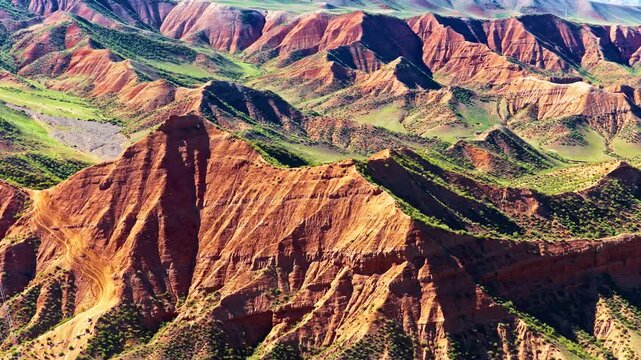 Aerial view of red Danxia landforms with steep eroded ridges, green vegetation, layered rock formations, and strong sunlight across rugged terrain.