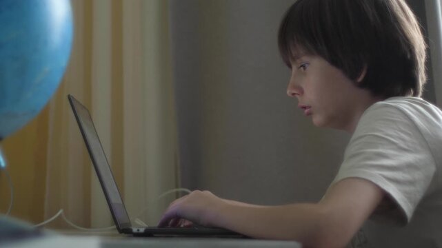 Boy focused on laptop while studying at home desk, remote education concept. Adolescent schoolboy doing homework online with a globe in the background