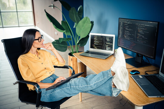 Female programmer relaxing with feet on desk while reviewing code on laptop and monitor in a modern home office, casual remote work and thoughtful software developer moment