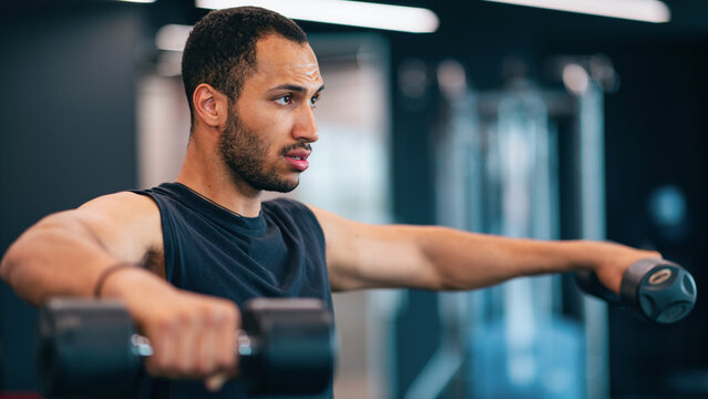 Focused Middle Eastern Man Performing Dumbbell Lateral Raises In Gym, Strength Training Session. He Is Engaged In Exercise Routine That Enhances Muscle Strength And Fitness.