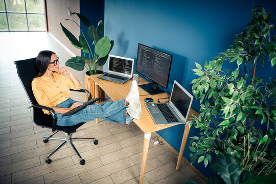 Relaxed woman programmer working remotely in a bright home office with laptops and monitors, feet propped on desk while reviewing code and planning development tasks