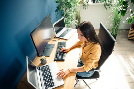 Woman working on multiple computers in a bright home office, remote work setup with laptop and monitor for programming and multitasking
