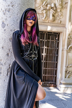 Woman wearing a dramatic black gothic costume with hood, corset, chains and decorative purple mask poses beside an ornate historic building, creating a mysterious dark fantasy portrait
