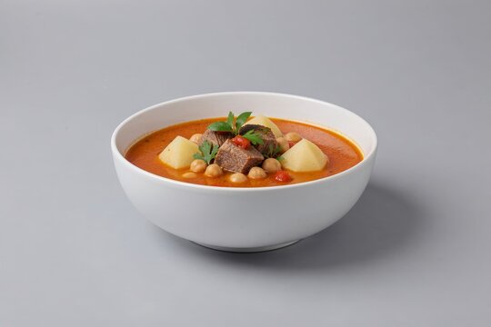 Bowl of Bozbash, vibrant lamb soup with chickpeas, potatoes, herbs. Rich orange color, high quality, plain grey background. Concept of hearty, comforting dish, ideal for food lovers.