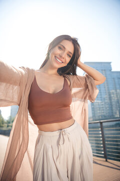 Smiling young woman taking a selfie on a sunny city boardwalk wearing casual summer outfit and lightweight cardigan