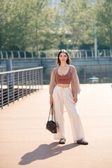 Fototapeta premium Young woman in casual summer fashion walking on urban boardwalk with handbag — relaxed confident portrait in sunny city park