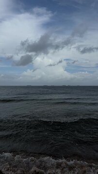 Dark ocean waves rolling toward a rocky shoreline beneath a cloudy blue-gray sky, with layered clouds and a distant horizon creating a natural coastal seascape.