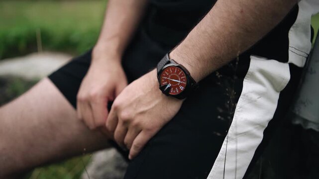 The man is sitting on a stone, and he has a watch on his wrist. The camera takes a close‑up shot.