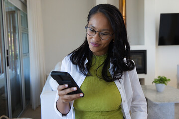 Obraz premium African American woman holding smartphone and looking down in living room wearing glasses olive top