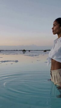 Vertical video: Shifting woman making ripples while seeing sunset in lagoon, tied shirt, copy space