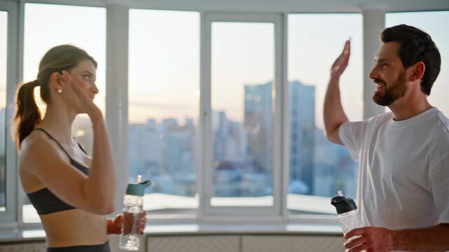 Couple high five celebrating home workout. Man drinking water panoramic window.
