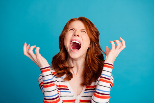 Angry frustrated woman shouting and screaming with hands raised &mdash; expressive portrait of a redheaded young woman in a striped sweater against a blue background
