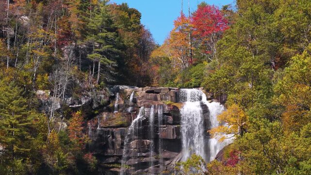 Majestic cascade pours through wooded mountain gorge surrounded by colorful autumn trees. Mist and rushing water create tranquil natural scenery.