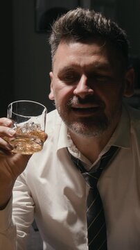 Vertical video of middle aged Caucasian man drinking whiskey from glass after work. Ideal for mental health campaigns, addiction recovery marketing, and workplace burnout awareness projects