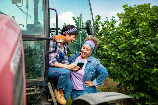 Two women farmers using smartphone by tractor in orchard