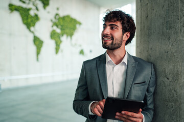 Fototapeta na wymiar Young businessman holding digital tablet, smiling and looking away, world map on wall, global business strategy