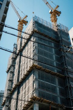 Tall building under construction with safety netting and cranes against a clear blue sky. Power lines cross in the foreground, suggesting an urban work zone.