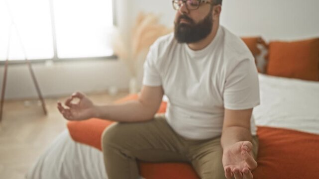 Man sitting on bed with hands in mudra and eyes closed, beard and glasses visible beside orange pillows and blanket in bedroom; serenity mindfulness.