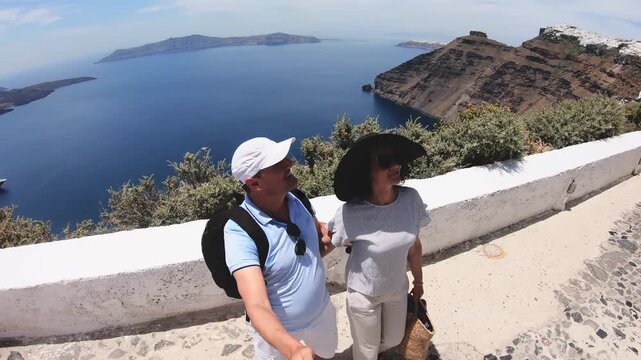 A man and woman are posing for a picture on a stone walkway on summer vacation trip in Santorini island in Greece overlooking the ocean. 
