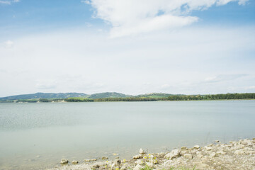 Tranquil Lake San Giuliano in Italy with calm waters, wooded shores, and distant hills under a cloudy sky. Rocky shoreline with wildflowers frames serene scenery for travel and nature themes. © Grenar