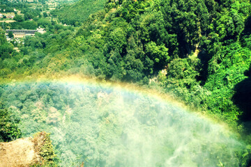 Vibrant rainbow arcs over misty forest valley at Marmore Falls, Umbria, Italy. Water spray rises through lush greenery creating magical atmosphere. Enchanting nature scene for travel themes. © Grenar