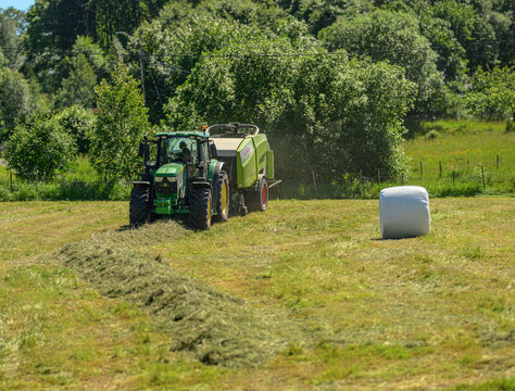Lindesnes, Norway - June 15 2026: Distant tractor and baler harvesting hay beside white bale.