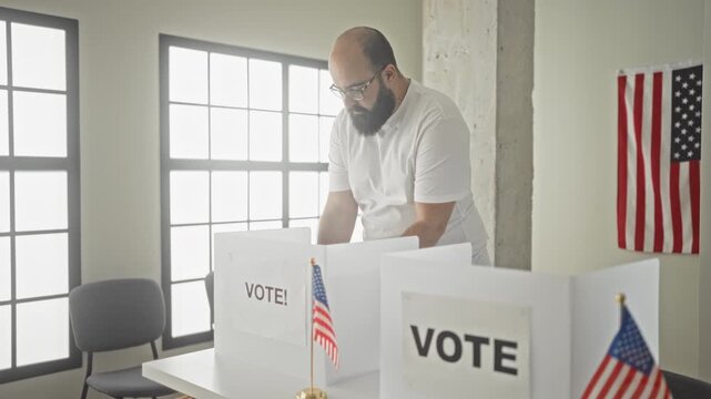 Man affixing sticker on shirt at a voting booth with small american flags and cardboard ballot screens in building; civic duty pride.
