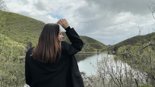 A woman putting glasses on standing on a hill overlooking a lake and mountain. The sky is cloudy and the water is calm