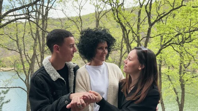 Happy family of a woman and two young kids are standing in a forest smiling, with the woman holding hands with one of the men